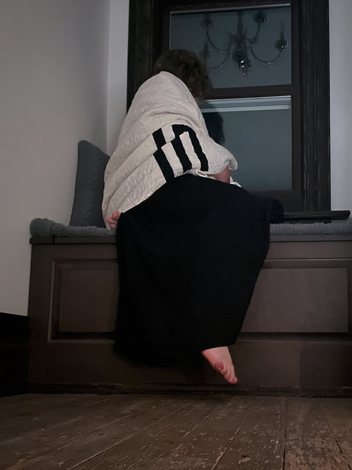 A person sits barefoot on a dark wooden window seat, wrapped in a cream knit sweater with black stripes, looking out a dark window in a dimly lit room with wooden floors.