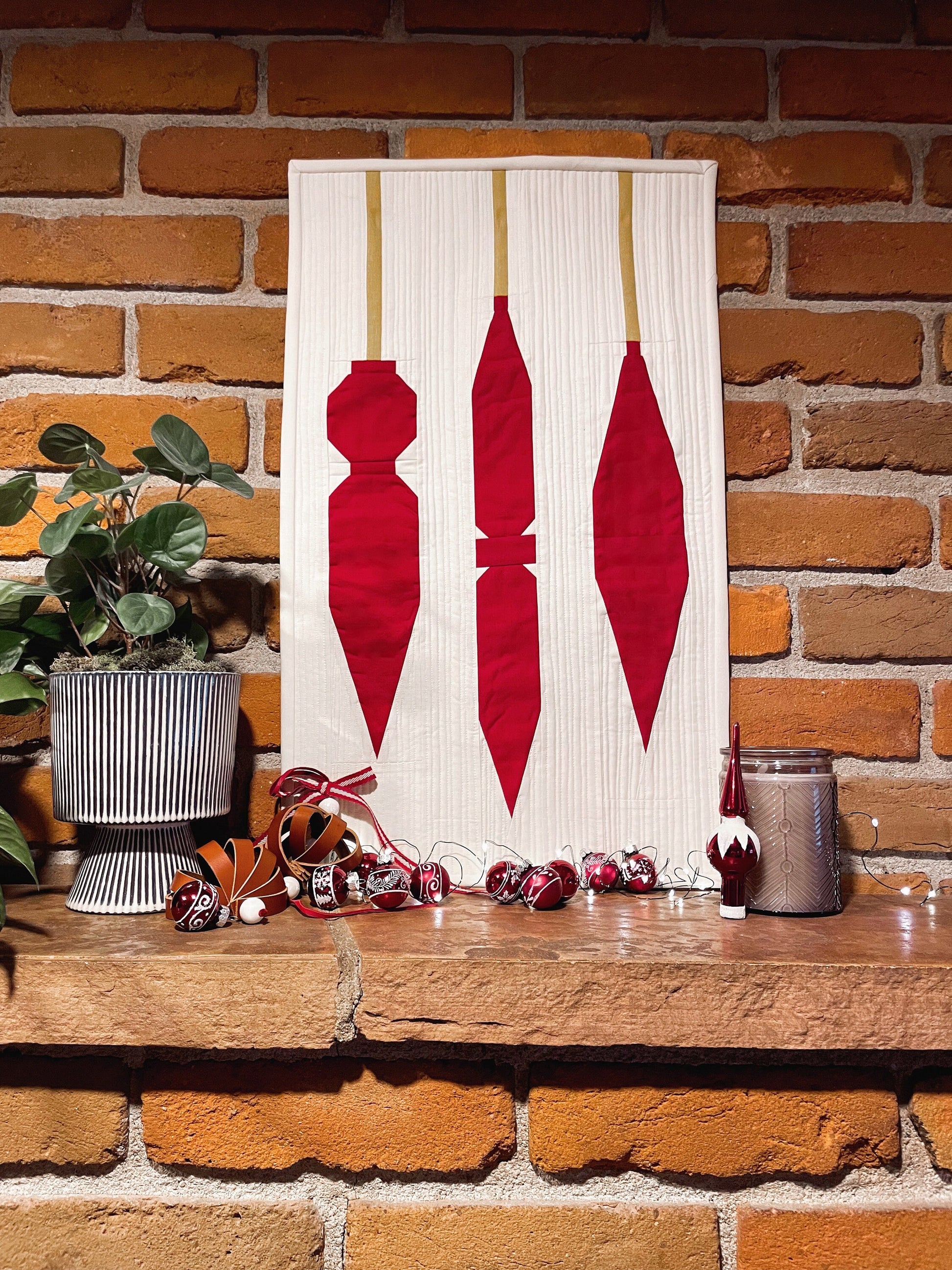 a modern quilt of three ornaments hangs on a brick wall with glass ornament balls in front of it. a plant is next to it. 