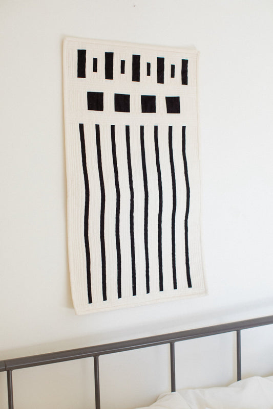 Side view of a modern minimalist quilt with a bed in the background. 