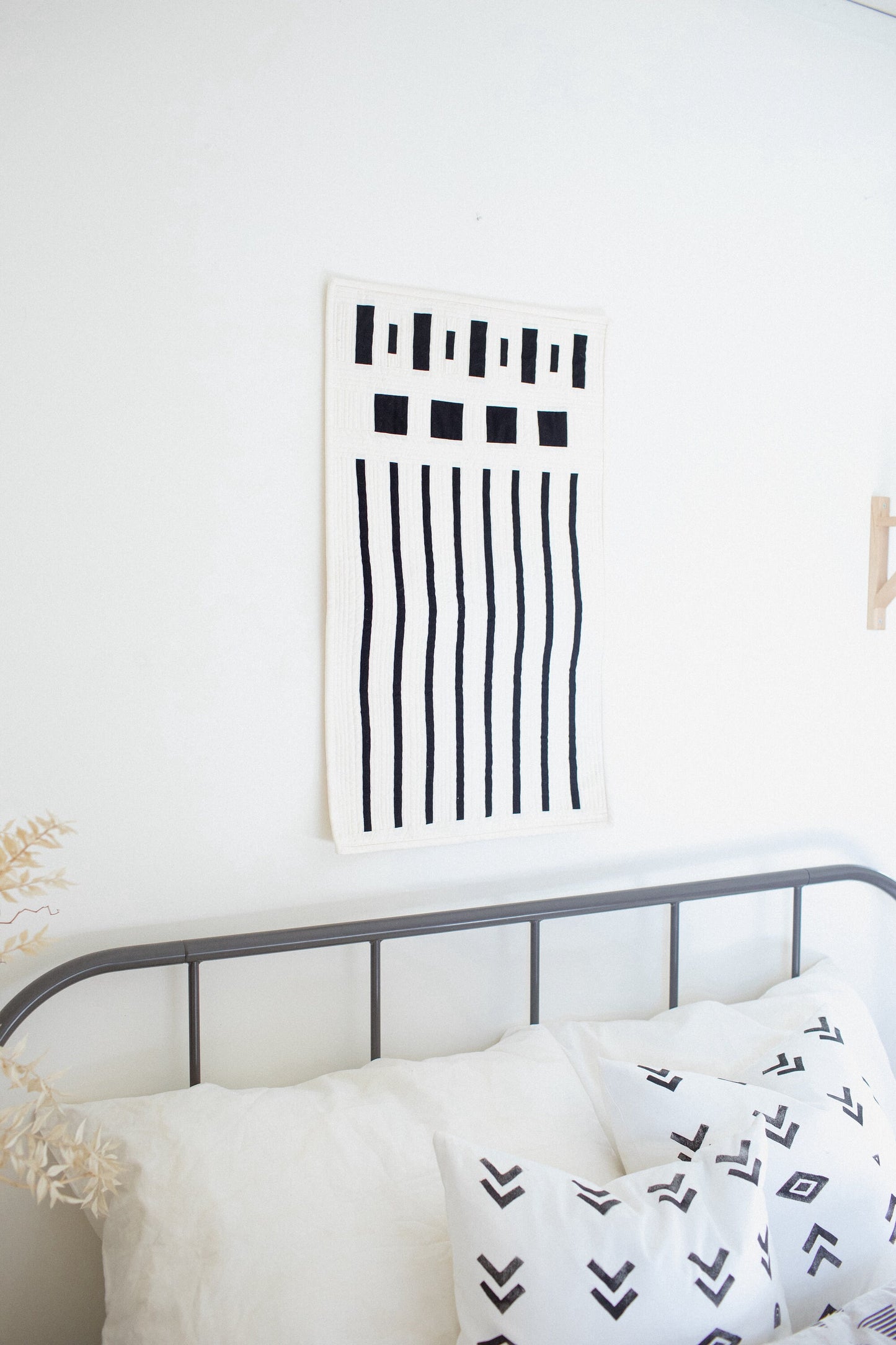 A modern quilt with a minimalist design featuring bold geometric lines displayed on a wall above a bed with white bedding.
