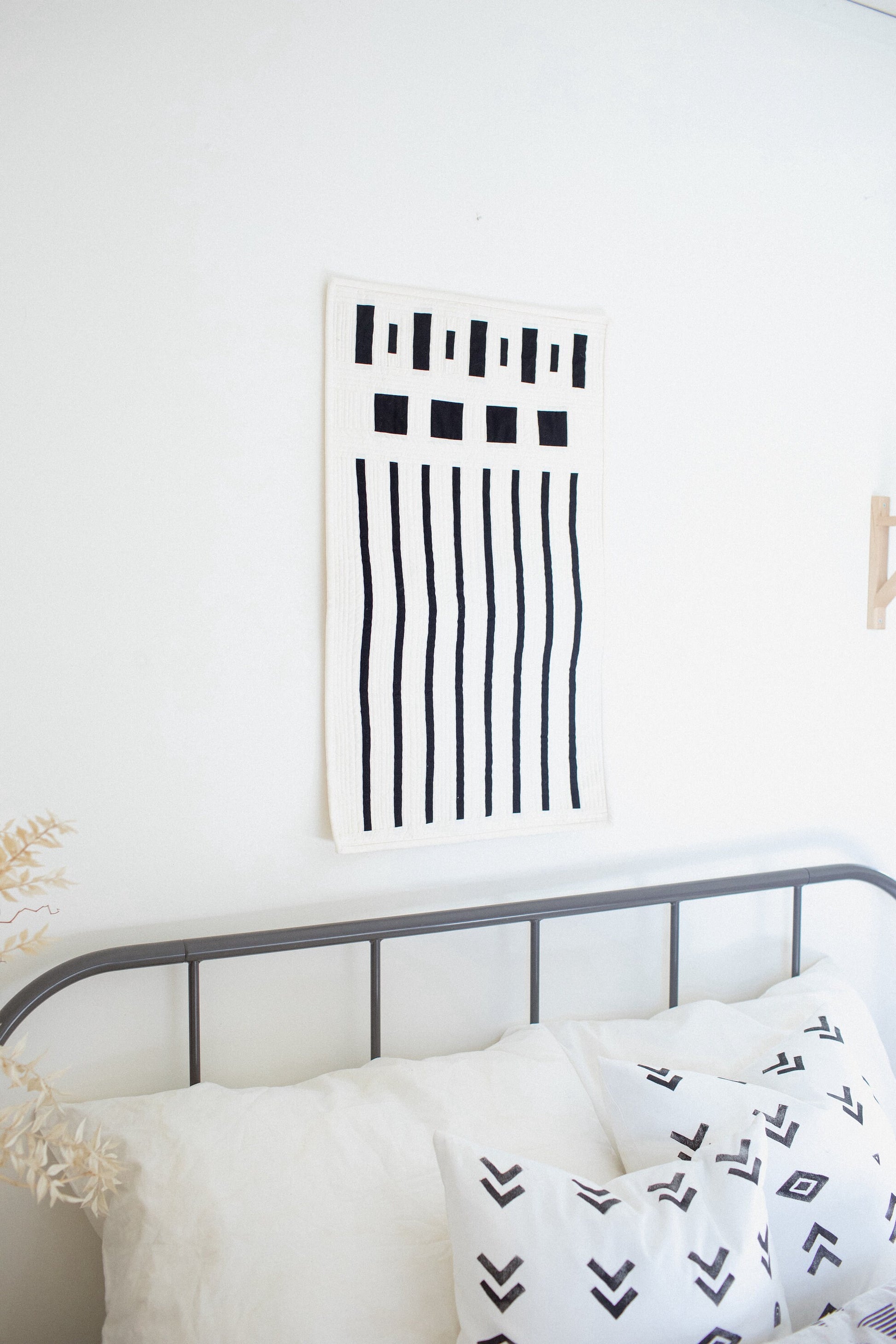 A modern quilt with a minimalist design featuring bold geometric lines displayed on a wall above a bed with white bedding.