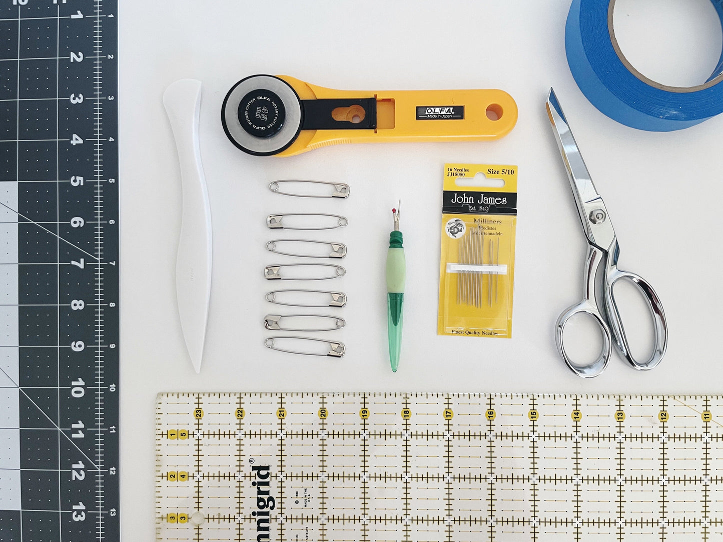 A flat lay of sewing supplies on a white surface including pins, needles, scissors, rotary cutter tape, hera marker, ruler. 
