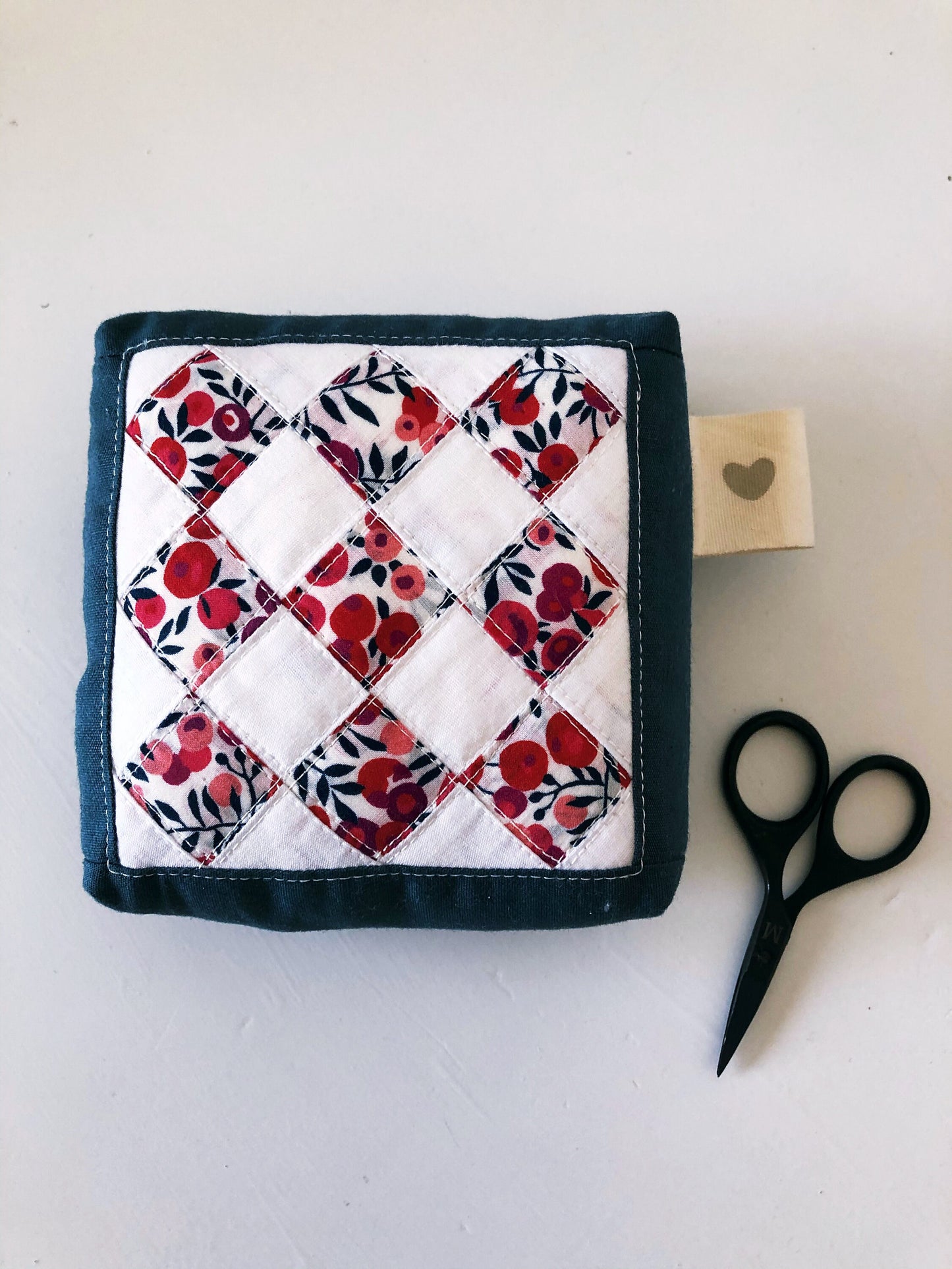 A top down view of a pincushions made of tiny scraps squares. a small pair of scissors lays next to it. 