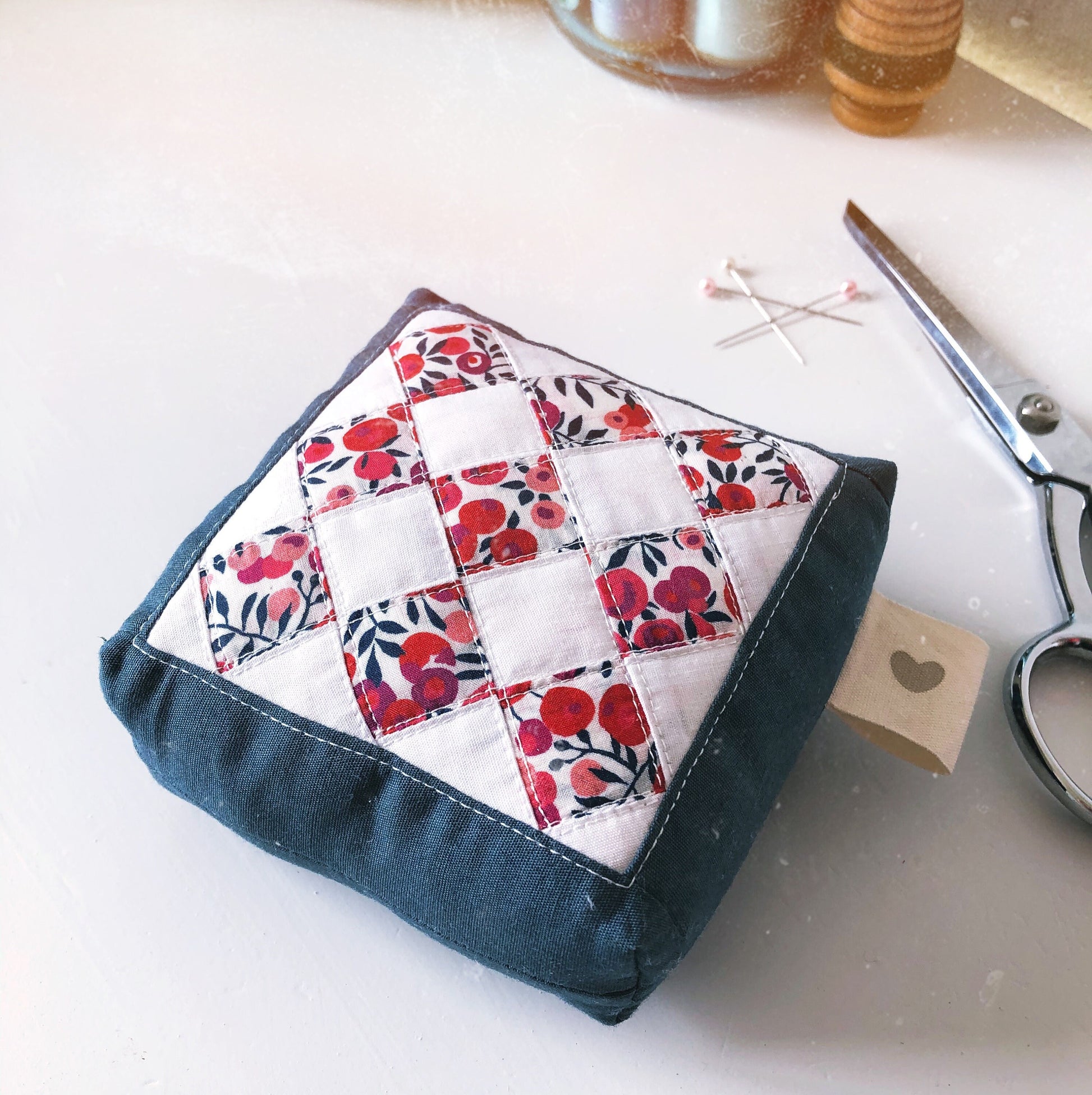 A quilted pincushion with a modern geometric design, featuring multicolored fabric patches, displayed next to a pair of scissors on a white surface.