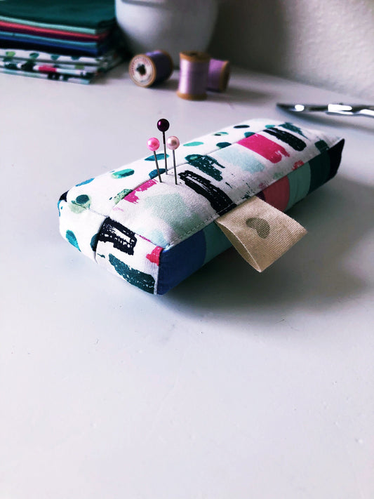 A rectangle pincushion with a patchwork design, featuring a variety of colors and patterns, placed on a table with sewing-related items in the background.