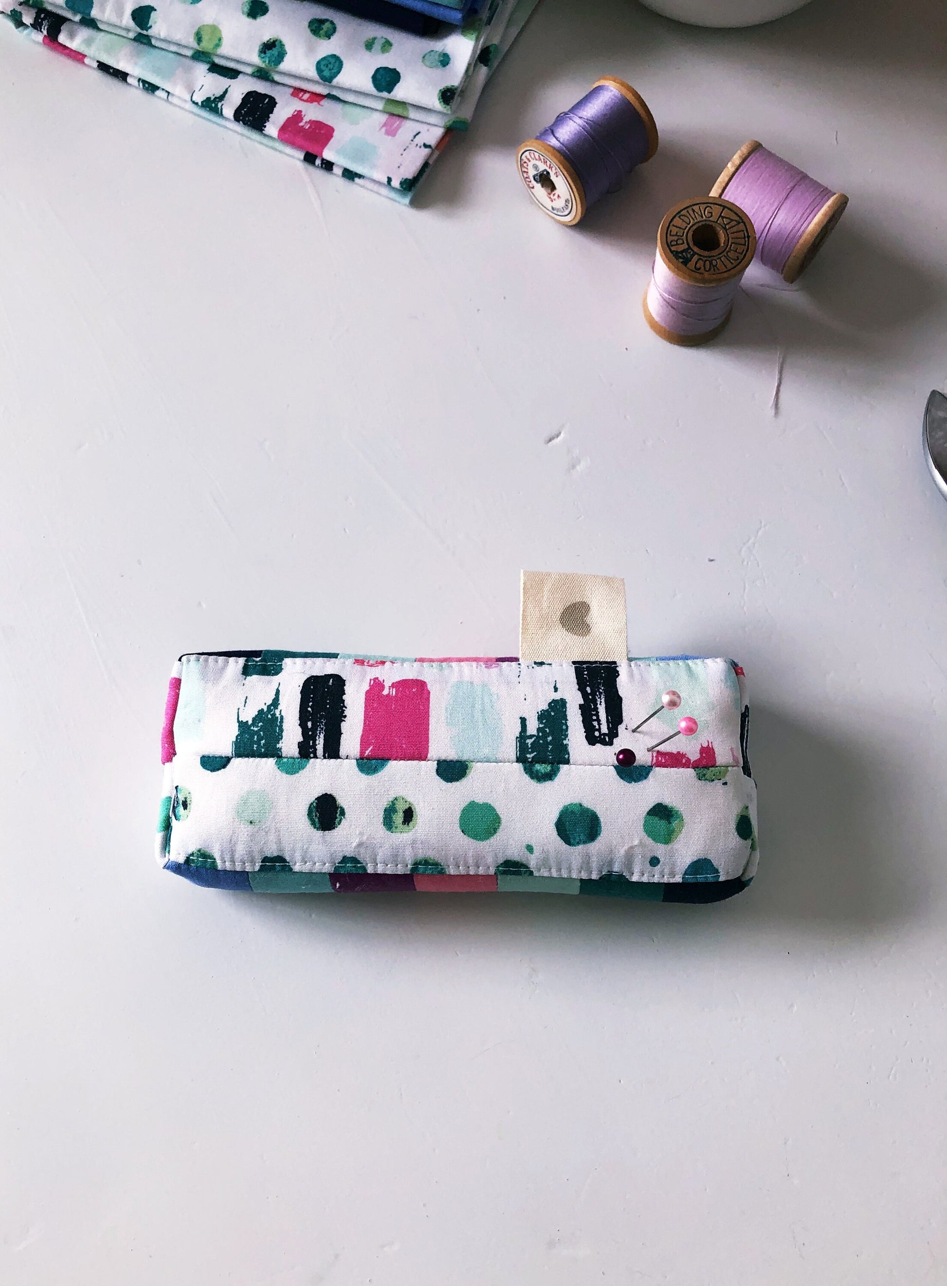 Top down view of a rectangle shaped pincushion with two pins. Sewing related items are in the background.