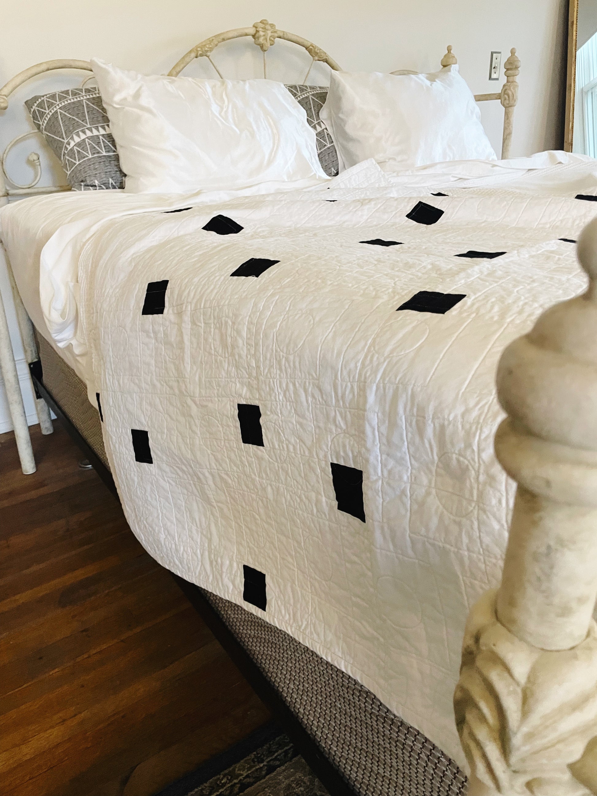Bed with white quilt featuring black diamond patterns, wooden floor, and decorative pillows.