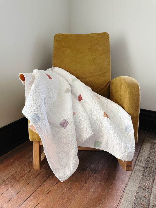 White quilt with colorful patterns draped over a mustard yellow armchair against a plain wall.