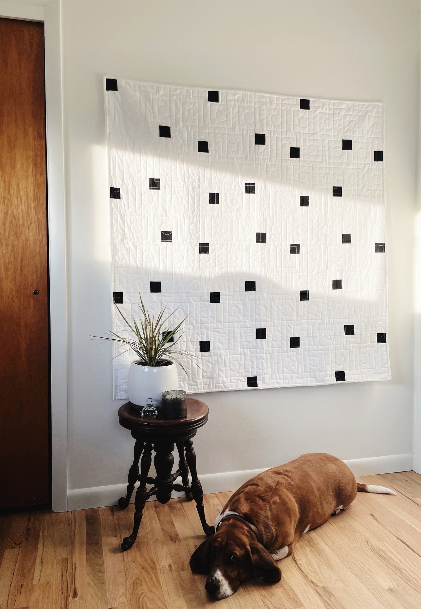 Modern geometric quilt with small black squares hangs on a wall with a basset hound sleeping on floor