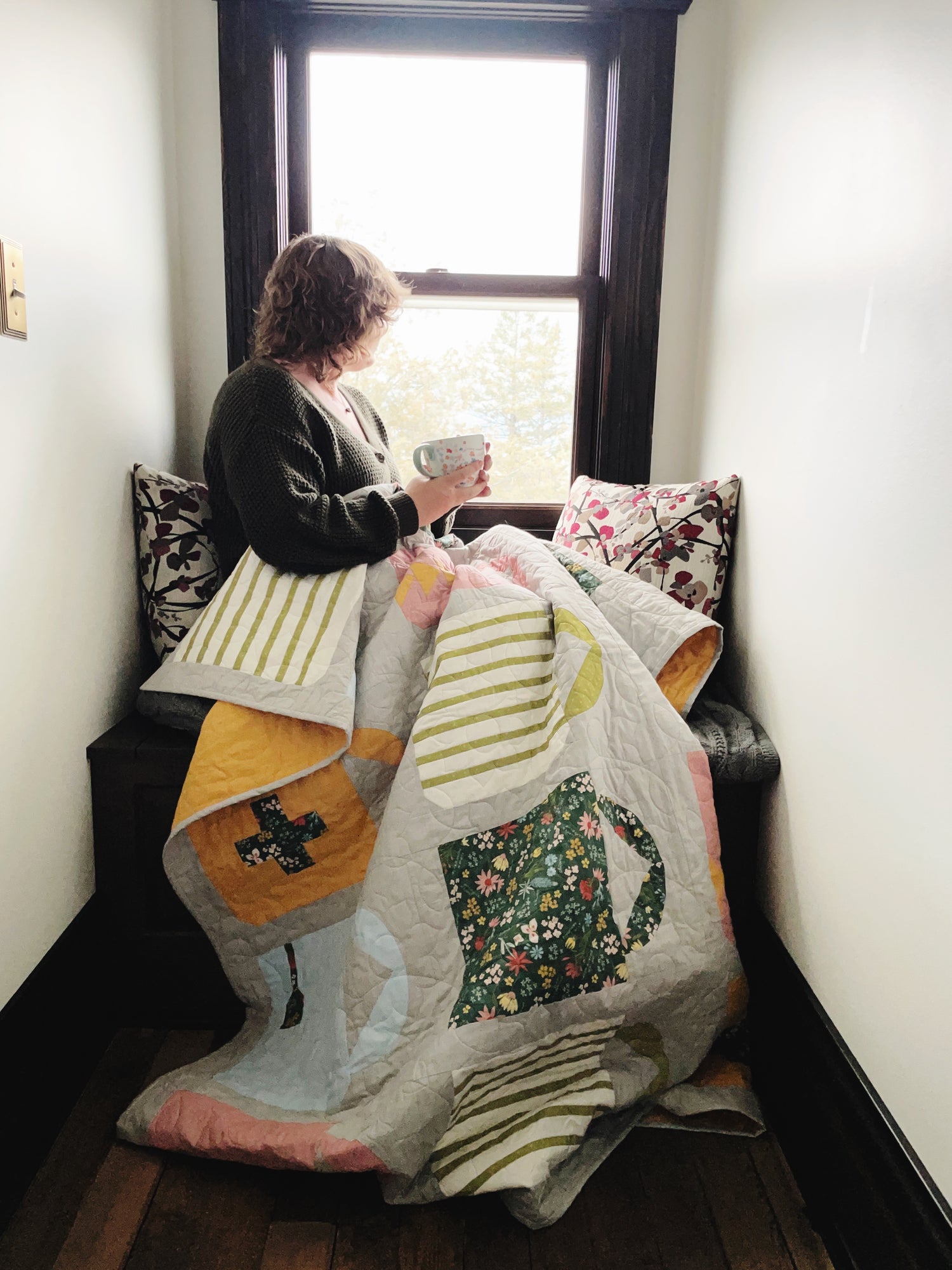 Person sitting on a bench with a colorful quilt draped over them, holding a cup.