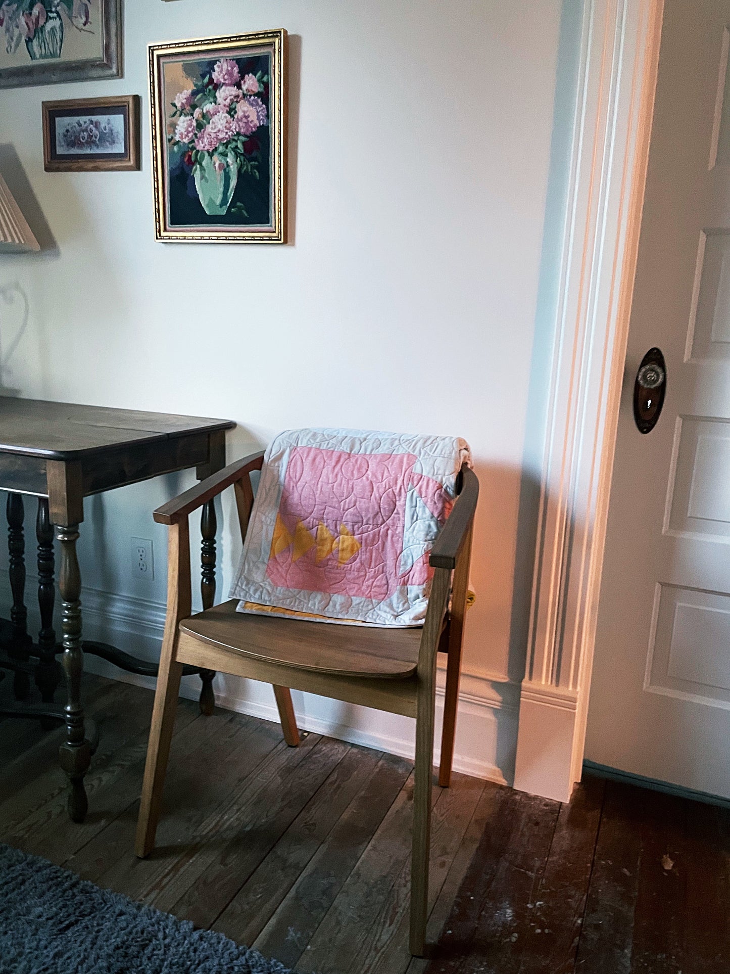 Chair with a pink mug quilt draped over it in a room with a table and framed pictures on the wall.