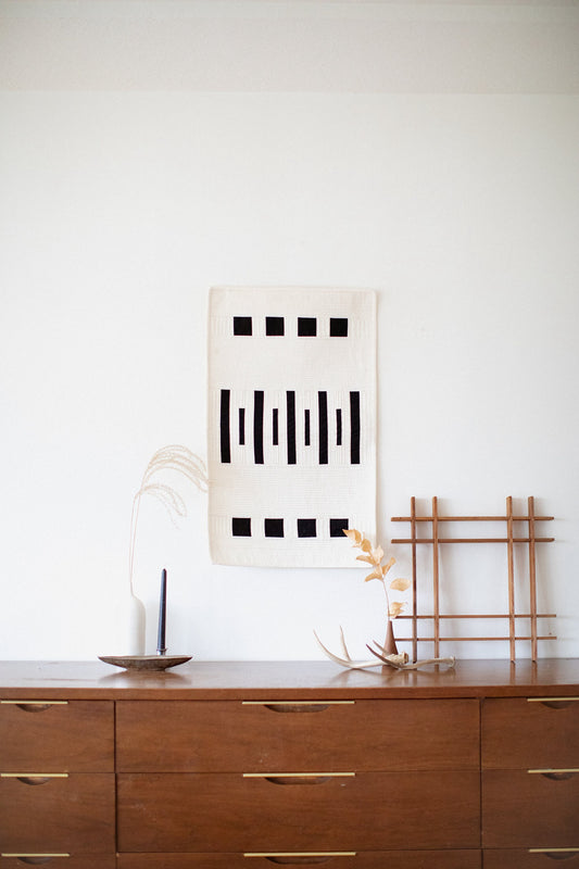 A quilt with a geometric pattern featuring aspen tree trunk motifs displayed on a wooden table alongside a candle and decorative items.