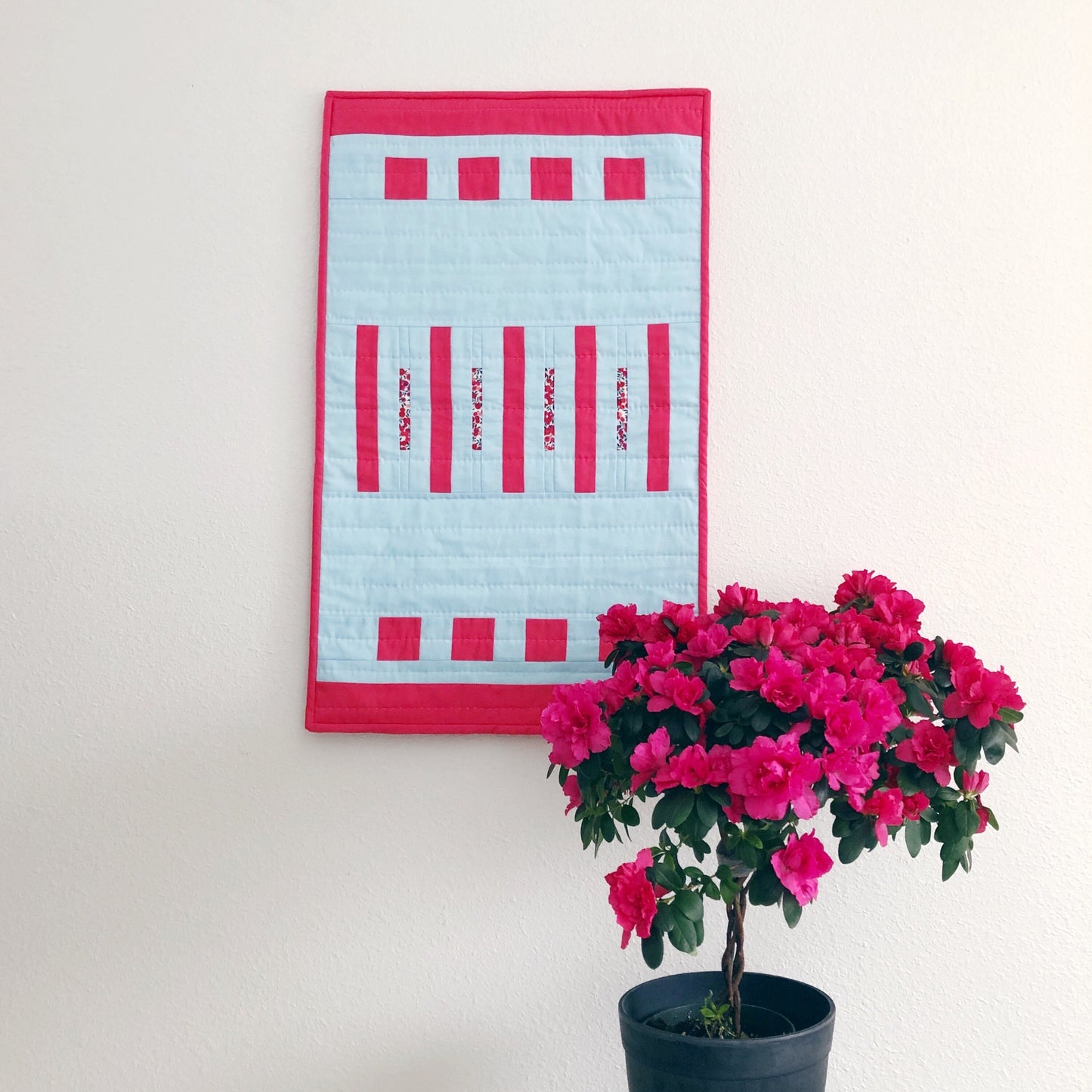 A modern minimalist Quilt with an azalea in front of it. 