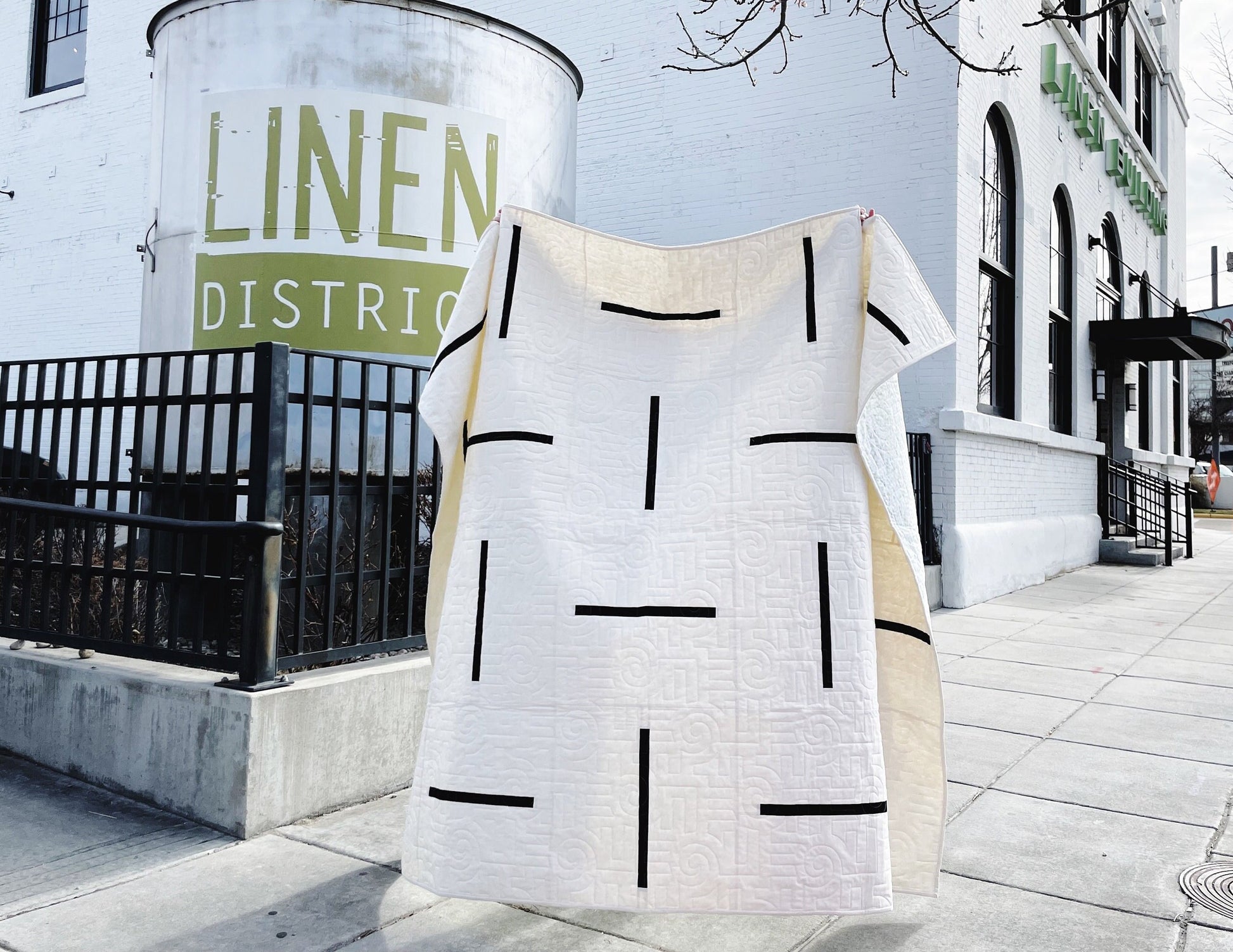 A quilt with modern geometric lines displayed in front of a building with 'LINEN DISTRICT' written on it.