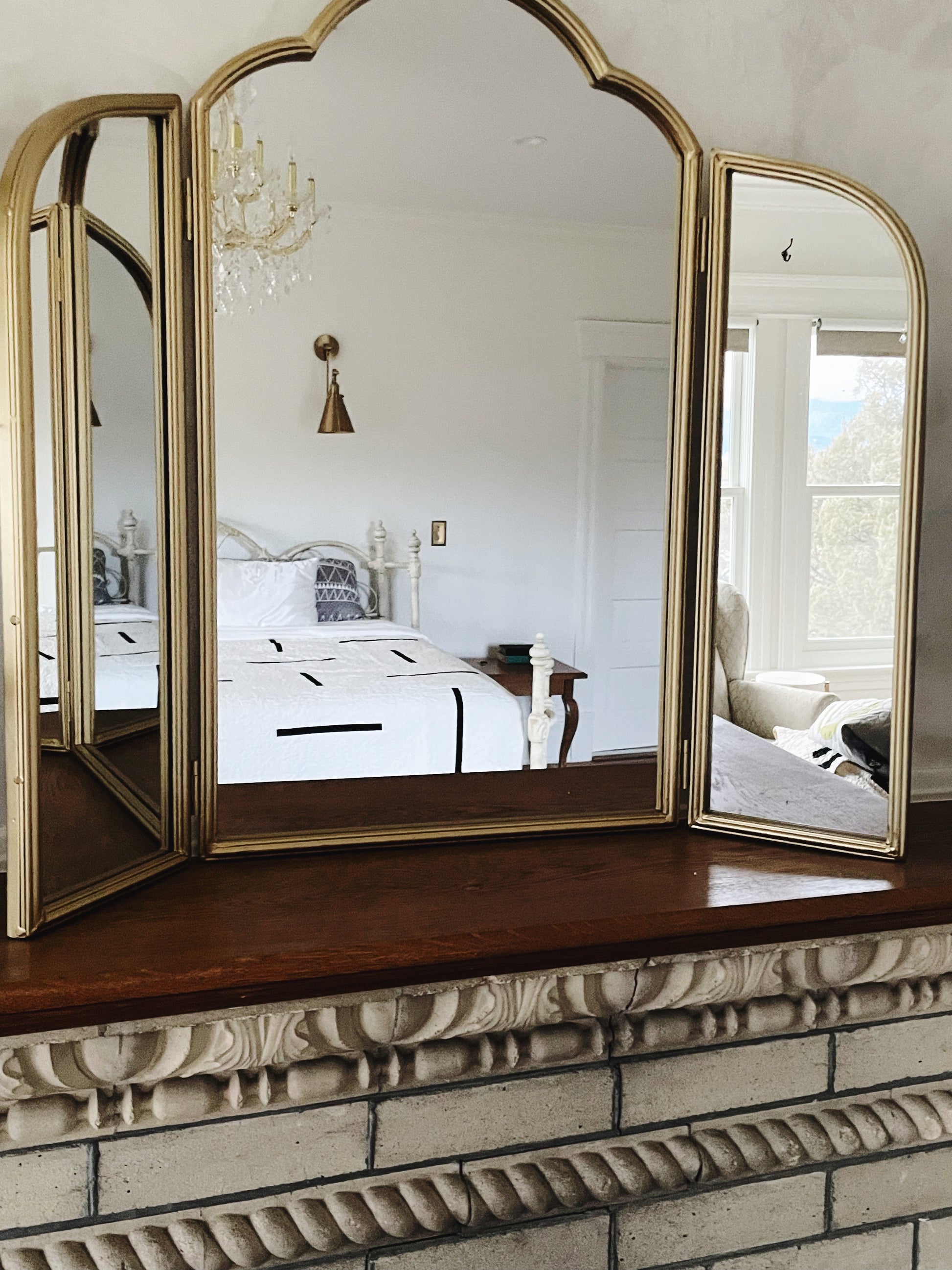 Tri-fold mirror on a decorative fireplace with a bedroom reflection.