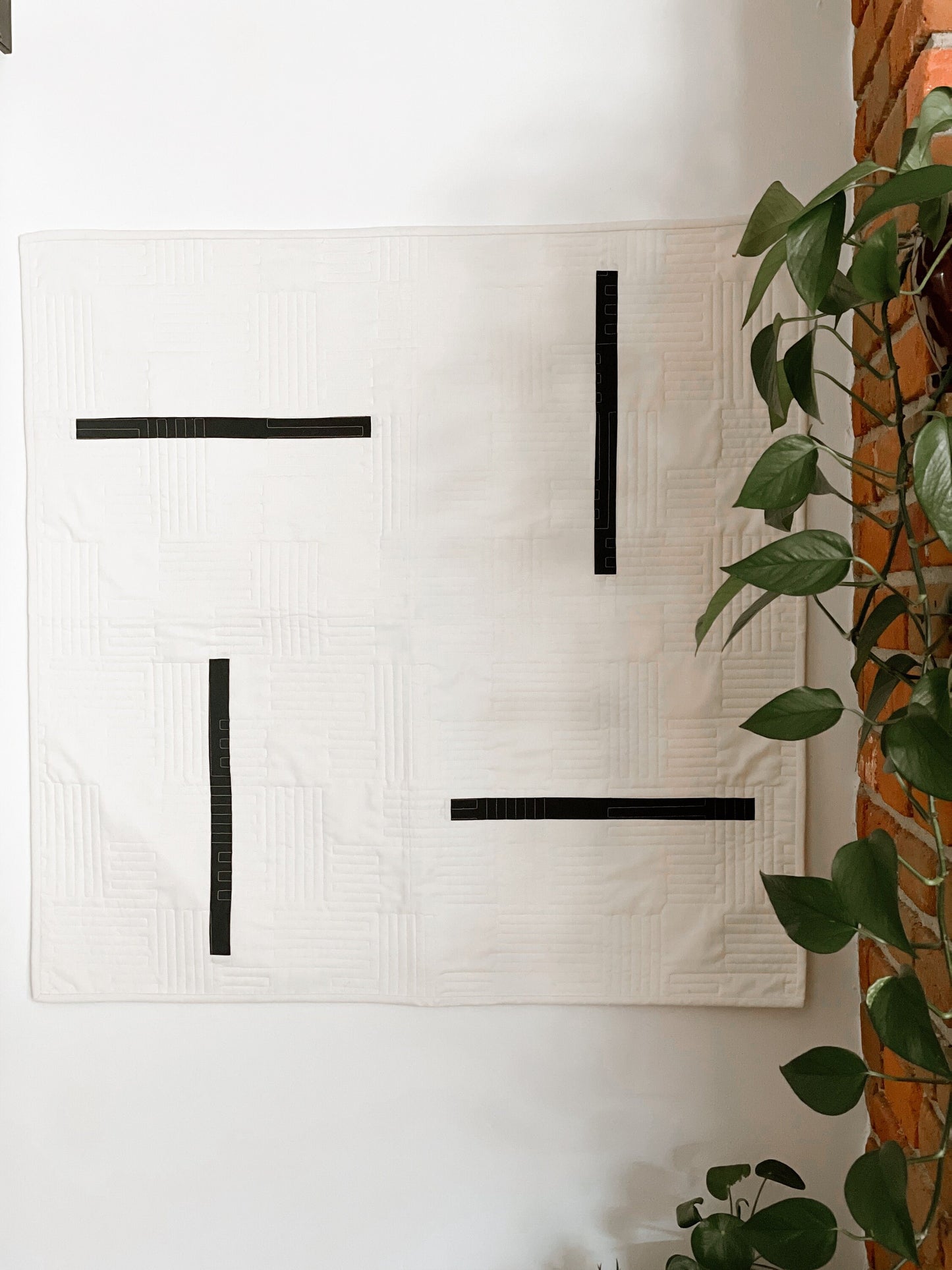 A modern geometric quilt with straight lines hangs on the wall next to a brick mantle with a plant on it. 