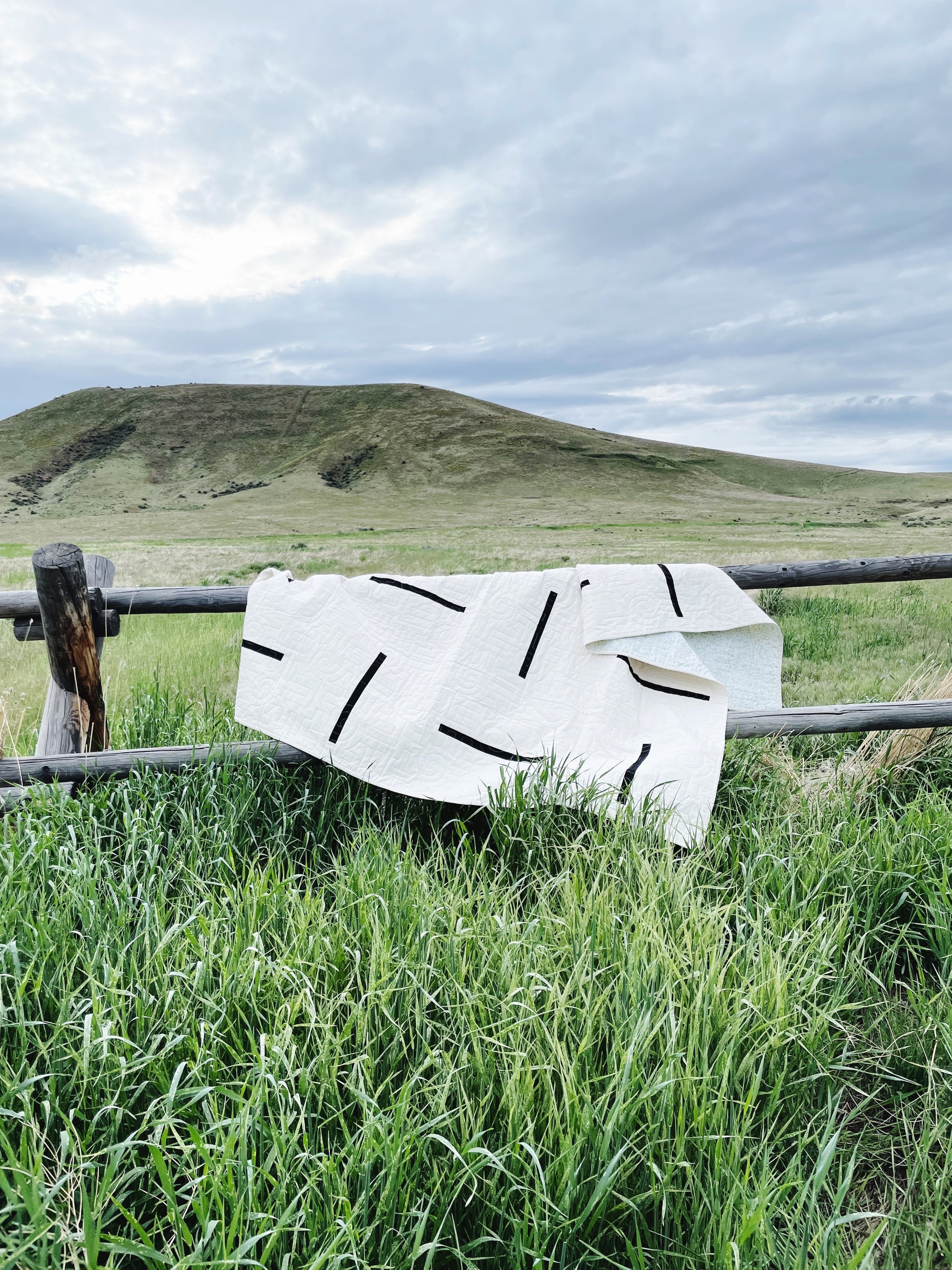 A modern minimalist quilt lays over a wooden rail fence with a hill in the background. 