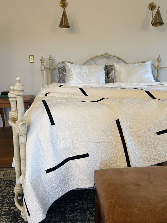 Bed with a white quilt featuring black geometric patterns, in a room with wooden floors and wall sconces.