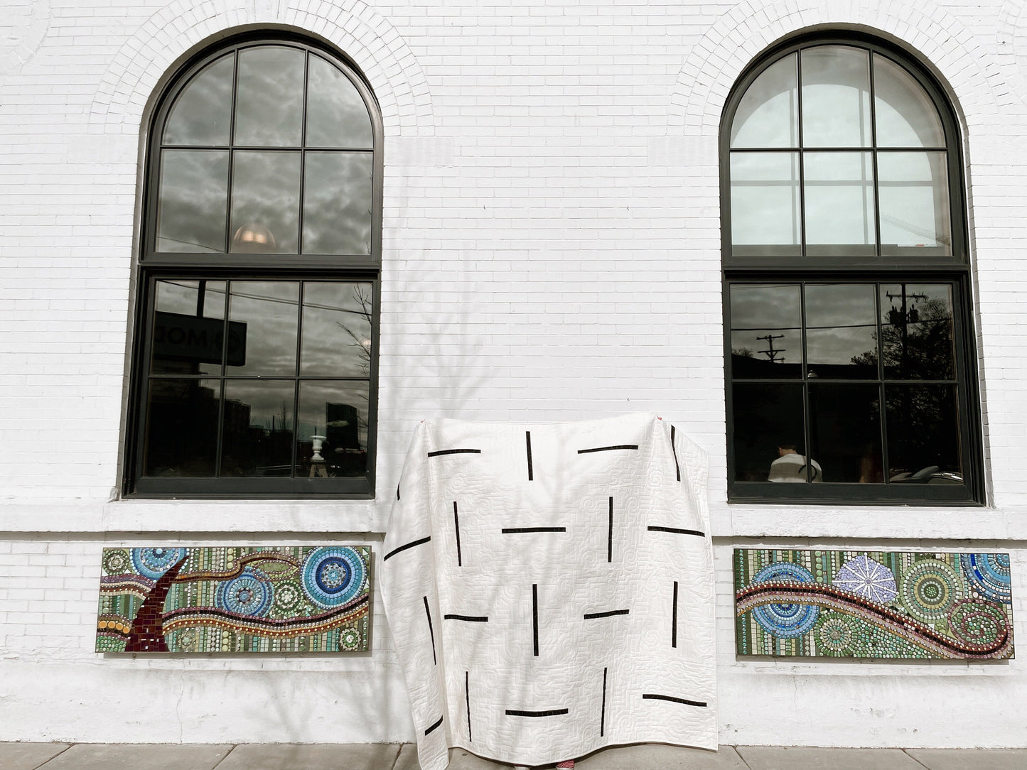 a modern geomtric quilt is on front of a white brick wall between two arched windows with mosaics underneath.
