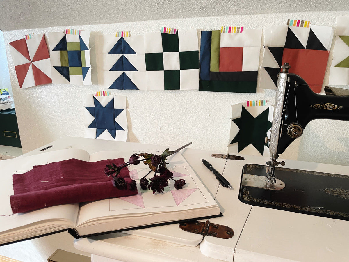 A quilt journal lays on a sewing desk next to a vintage sewing machine. A flower and matching fabric are open to a page with a quilt block drawn on it. Quilt blocks are hanging on the wall in the background. 