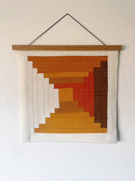 Quilted log cabin  wall art with geometric pattern in earthy tones hanging on a white wall.