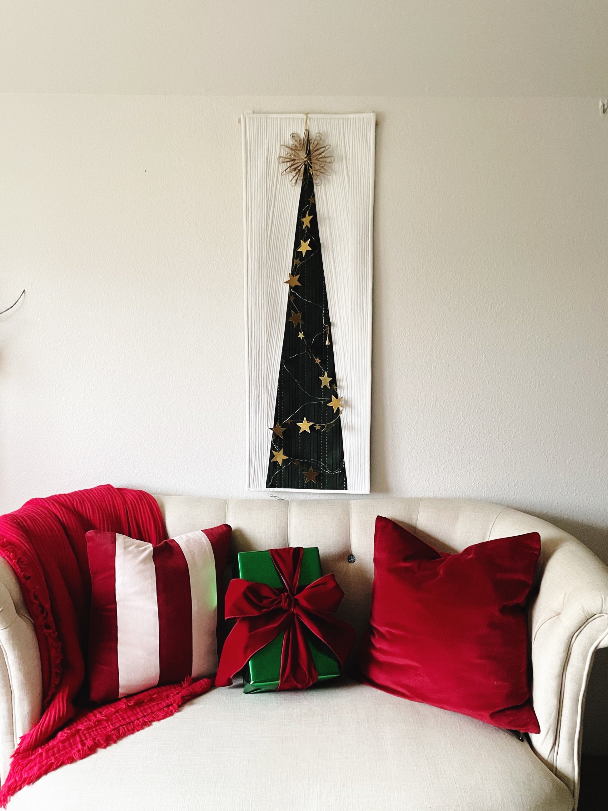 Decorative Christmas tree wall art above a sofa with red and green pillows and a gift.