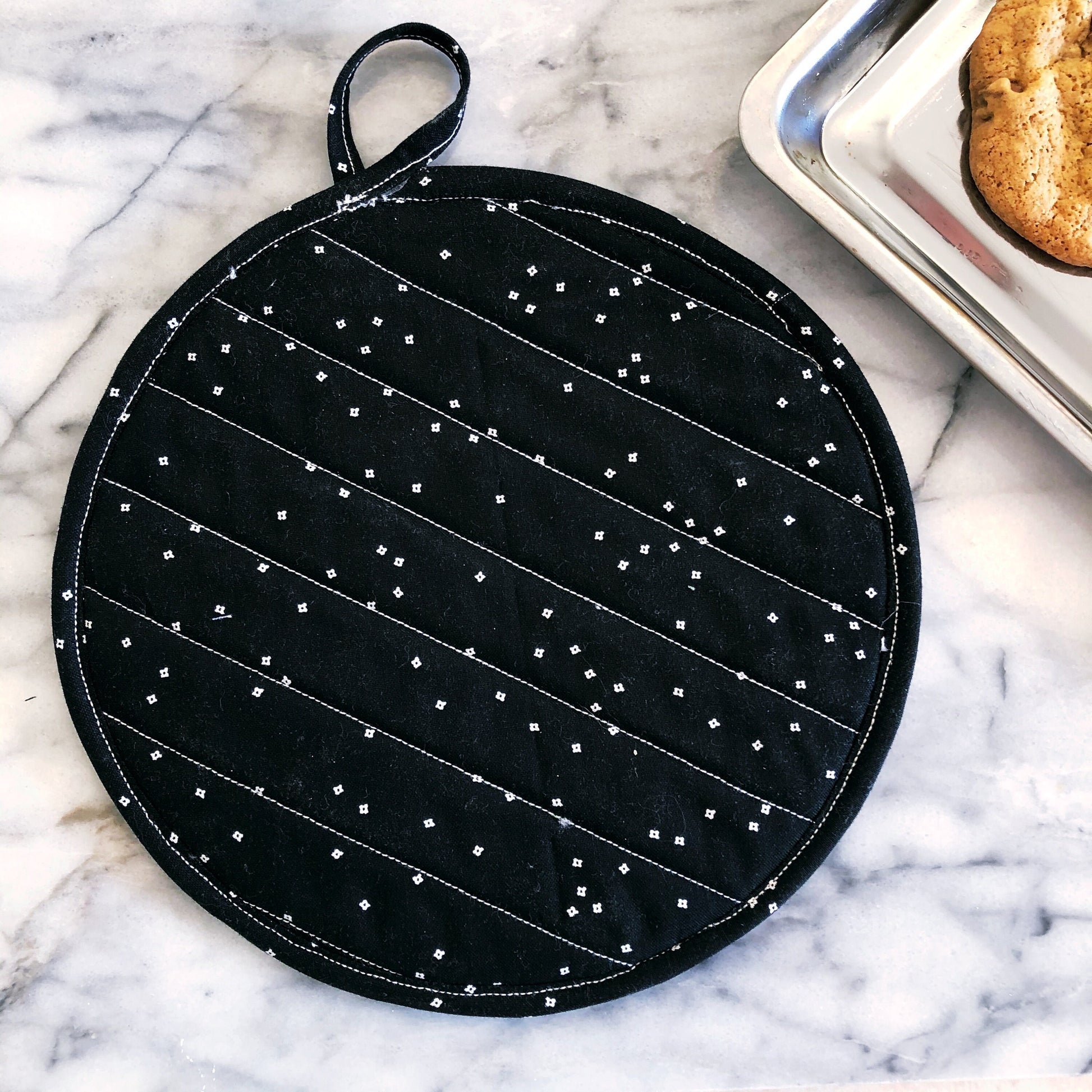 a pot holder with a tab lays on the counter. Cookies can bee seen in the background. 