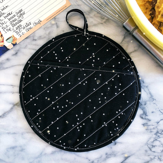 A black fabric pot holder with a geometric pattern and a lanyard for hanging, displayed on a marble surface.