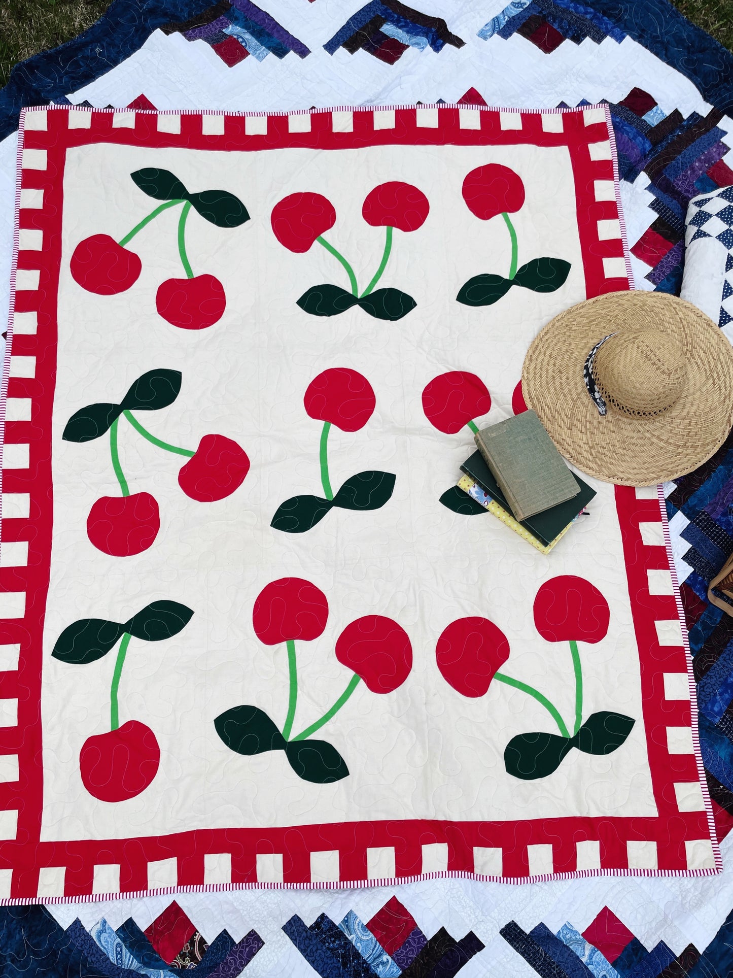 A top down view of a bright cherry quilt laying on the ground. Books and a straw hat are laying on it suggesting a picnic