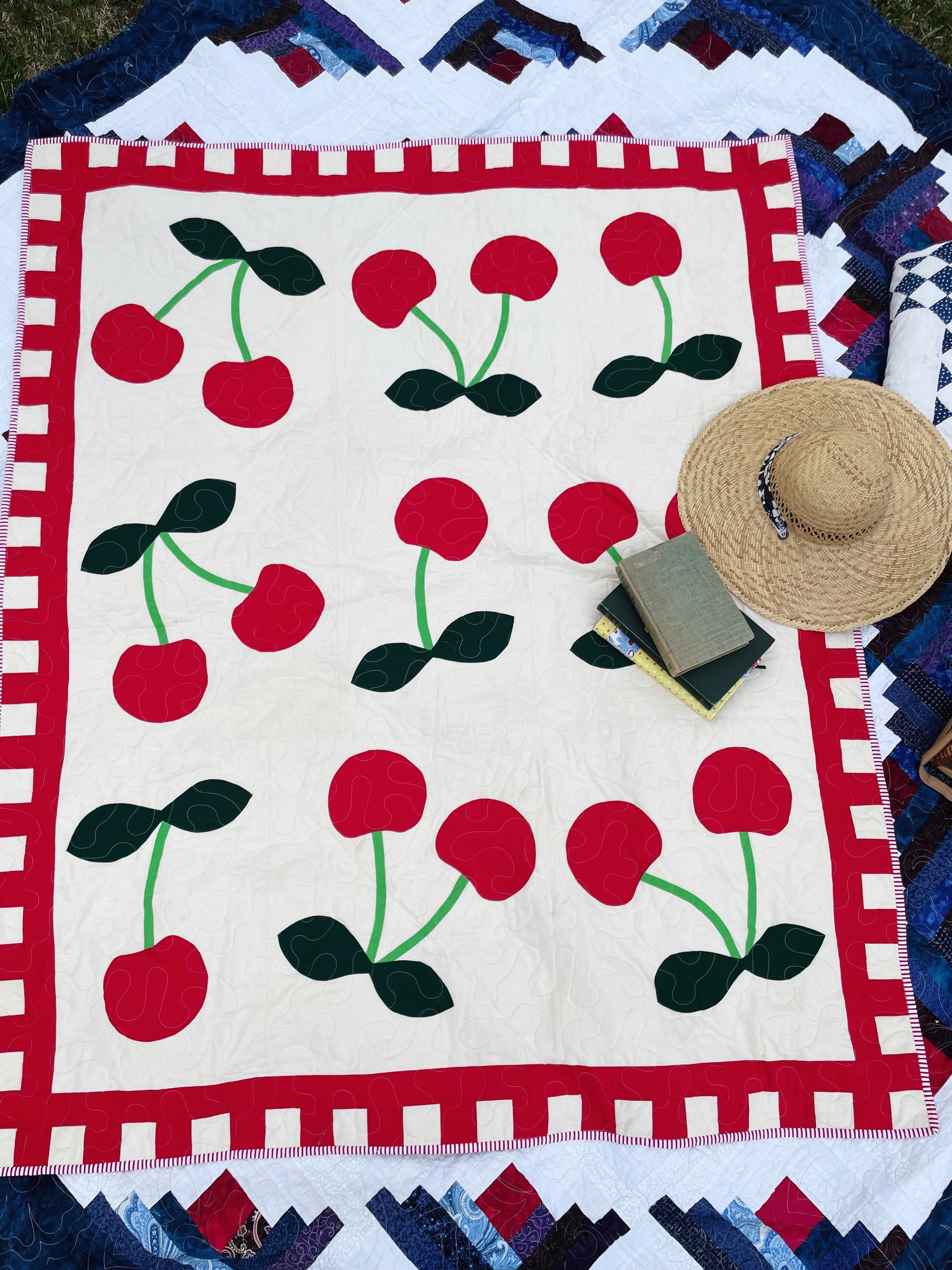 A top down view of a bright cherry quilt laying on the ground. Books and a straw hat are laying on it suggesting a picnic