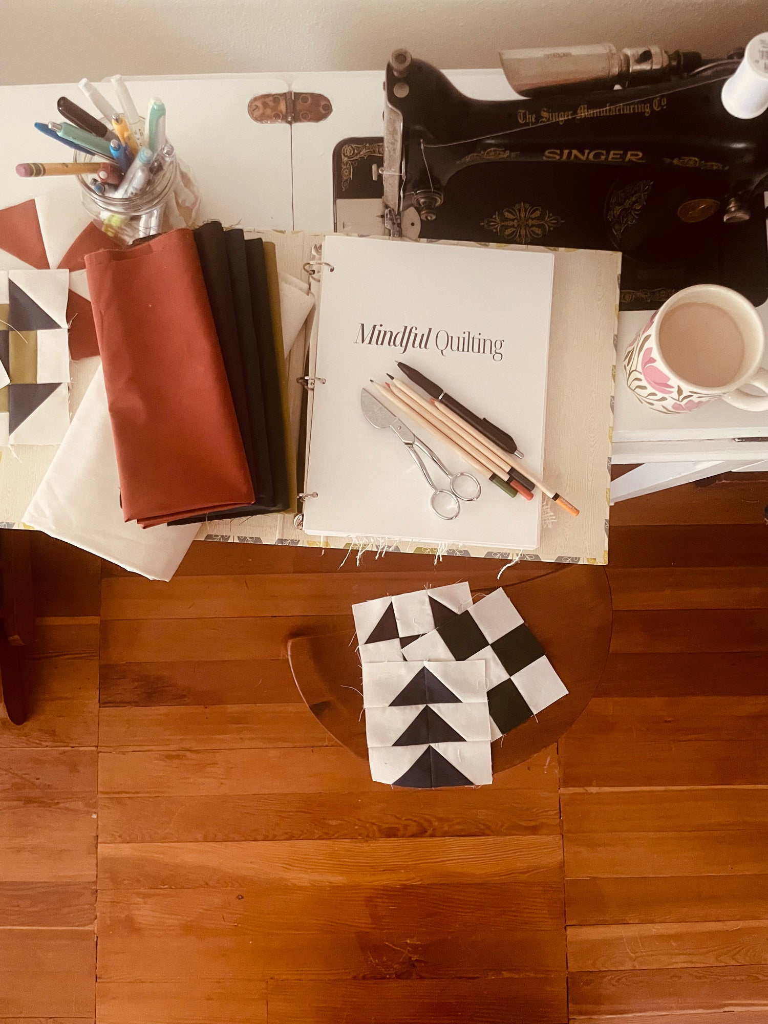 A top down view of a Mindful quilting journal open on a sewing desk. A sewing machine, sewing tools, fabric and quilt blocks are scattered around.