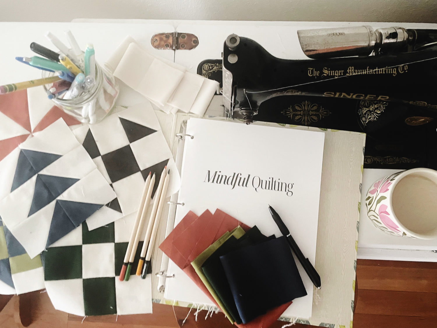 A top view of a quilt journal called Mindful Quilting lays on a desk with pencils, fabric and quilt blocks laying around 